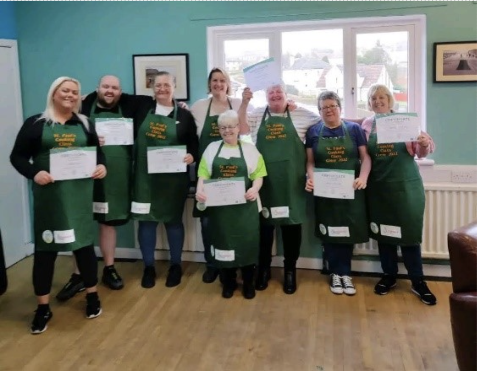 A group photo of the cooking crew wearing their aprons and holding certificates. All the people in the photo have a light skin tone. A group photo of the cooking crew wearing their aprons and holding certificates. All the people in the photo have a light skin tone.